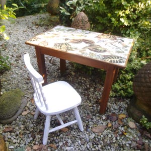 May include: A white wooden chair sits next to a small table with a patterned tabletop. The table and chair are outdoors on a gravel surface.