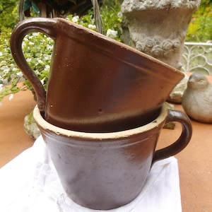 May include: Two brown ceramic mugs with handles. The mugs are stacked on top of each other. The top mug is slightly larger than the bottom mug.