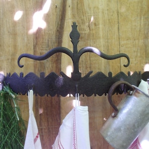 Könnte beinhalten: Ein schwarzer Wandhalter aus Metall mit mehreren Haken, der ein dekoratives Design aufweist. Der Halter hält ein weißes Tuch, eine grüne Pflanze und einen silbernen Krug.