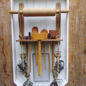May include: White wooden rack with a rolling pin and various wooden kitchen utensils, including spoons, spatulas, and a rolling pin. The rack is mounted on a wooden wall.