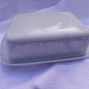 Puede incluir: Plato de mantequilla de porcelana blanca con un diseño floral gris. El plato tiene una tapa y una base.