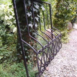 Puede incluir: Una valla de metal negro con un diseño decorativo, parcialmente oculta por vegetación verde. La valla está hecha de hierro forjado y tiene una forma curva.