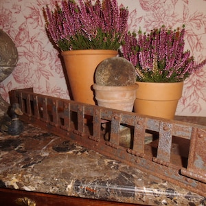 Puede incluir: Un objeto metálico rectangular y oxidado con aberturas rectangulares, posiblemente una rejilla decorativa. Dos macetas de terracota con plantas de flores moradas y un pequeño objeto redondo cubierto de musgo están en el fondo. También hay una figura de pato.