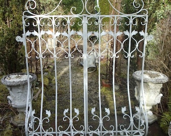 May include: A white wrought iron garden gate with floral and scrollwork details. The gate is flanked by two stone urns and set against a backdrop of greenery. The gate features a flower at the top center.