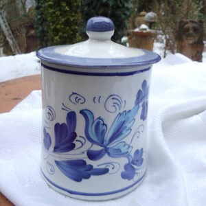 Könnte beinhalten: Ein weißer Keramiktopf mit Deckel, verziert mit blauen Blumenmustern. Der Topf hat eine zylindrische Form und einen abgerundeten Deckel mit einem kleinen Knauf. Das Design zeigt stilisierte blaue Blumen und Blätter.