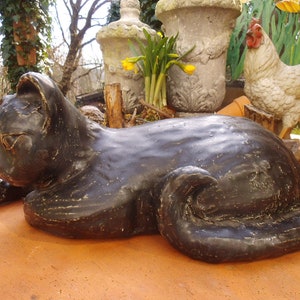 Może przedstawiać: Czarna ceramiczna figurka kota o gładkim, eleganckim wykończeniu. Kot leży z głową opartą na łapach. Figurka jest ozdobą do ogrodu lub domu.
