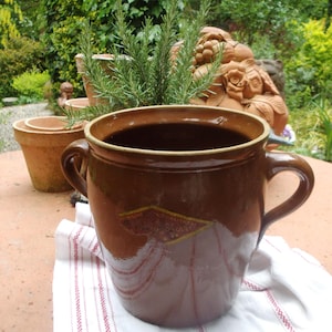 Op de afbeelding: Een grote, bruine keramische pot met twee handvatten, met een decoratief ruitvormig embleem. De pot staat op een wit en rood gestreepte doek, met een achtergrond van groen en andere terracotta potten. Een takje rozemarijn is zichtbaar.
