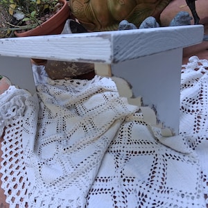 Può includere: Una mensola da parete in legno bianco con una superficie superiore rettangolare e due supporti laterali decorativi con finitura usurata. La mensola presenta una venatura del legno visibile e bordi consumati, mostrando un aspetto invecchiato.