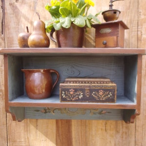 Op de afbeelding: Een houten plank met een grijs geverfd oppervlak en decoratieve snijwerk. De plank is gevuld met verschillende items, waaronder een bruine keramische kan, een kleine houten doos met bloemenontwerpen en twee peren. Een plant in een pot met gele bloemen staat op de plank.