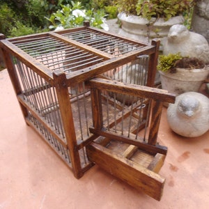 Puede incluir: Una jaula de pájaros de madera con una puerta de alambre de metal y un cajón extraíble. La jaula es de color marrón y tiene un aspecto rústico. Está sentada en una mesa con otros artículos decorativos.