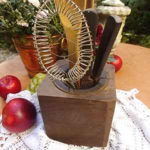 May include: A wooden utensil holder with a wire whisk, wooden spoons, and metal utensils. The holder is sitting on a white crocheted tablecloth with red apples.