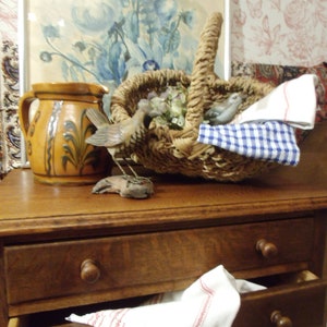 May include: A wooden two-drawer nightstand with a white cloth napkin hanging out of the top drawer. A brown ceramic pitcher and a wicker basket with dried flowers and two small bird figurines are on top of the nightstand.