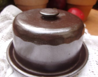 Può includere: Una campana per formaggio in ceramica marrone con un coperchio. La campana è appoggiata su un tovagliolo di pizzo bianco.