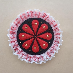 May include: A circular brooch featuring a black felt base with a red eight-petaled flower design. The center is accented with small gold-toned studs and beads. The entire piece is framed by a ruffled white lace trim along the outer edge.