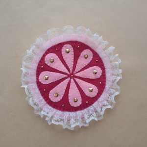May include: A circular felt applique featuring a deep magenta center with eight light pink petal-shaped segments arranged in a radial pattern. The piece is accented with small gold-toned beads and finished with a ruffled white lace trim border.