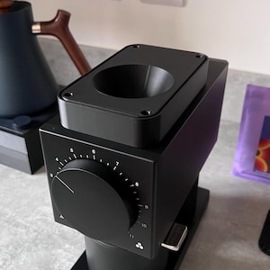 Könnte beinhalten: Eine schwarze elektrische Kaffeemühle mit einem silbernen Knopf und einem Zifferblatt mit den Zahlen 3 bis 11. Die Mühle hat einen schwarzen Trichter und einen schwarzen Auffangbehälter.