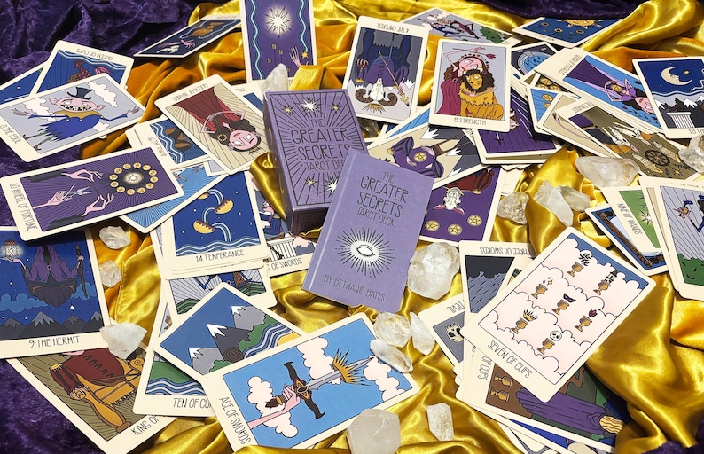 May include: A deck of The Greater Secrets tarot cards featuring purple and gold boxes, surrounded by numerous illustrated tarot cards with various figures and symbols, all resting on a draped gold and purple fabric surface with clear crystals.