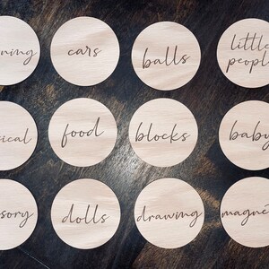May include: Twelve wooden circles with different words written on them. The words are: learning, cars, balls, little people, musical, food, blocks, baby, sensory, dolls, drawing, magnetic.