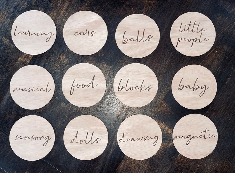 May include: Twelve wooden circles with different words written on them. The words are: learning, cars, balls, little people, musical, food, blocks, baby, sensory, dolls, drawing, magnetic.