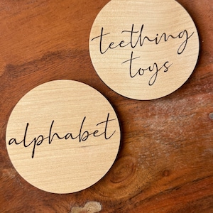 May include: Two round wooden signs with black lettering. One sign says "teething toys" and the other says "alphabet".