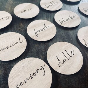 May include: A set of ten wooden circles with different categories written on them in black script. The categories include "cars", "balls", "blocks", "food", "dolls", "sensory", "musical", "little people", and two others that are not fully visible in the image.