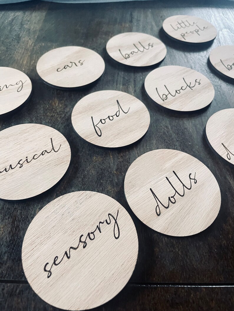 May include: A set of ten wooden circles with different categories written on them in black script. The categories include "cars", "balls", "blocks", "food", "dolls", "sensory", "musical", "little people", and two others that are not fully visible in the image.