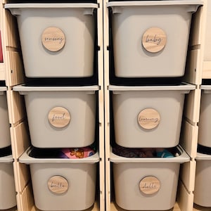 May include: A wooden shelf unit with six gray storage bins. Each bin has a round wooden label with a different word written on it: sensory, baby, food, learning, balls, and dolls.