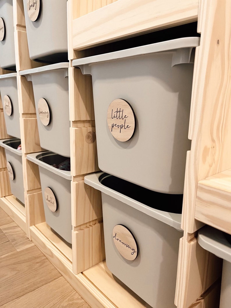 May include: A set of gray plastic storage bins with wooden labels. The labels are round and have the words "baby", "learning", "dolls", "little people", and "drawing" written on them.