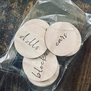May include: A set of six wooden round labels with handwritten text. The labels are in a clear plastic bag. The labels say "dolls", "cars", "blocks", "books", "games", and "puzzles".