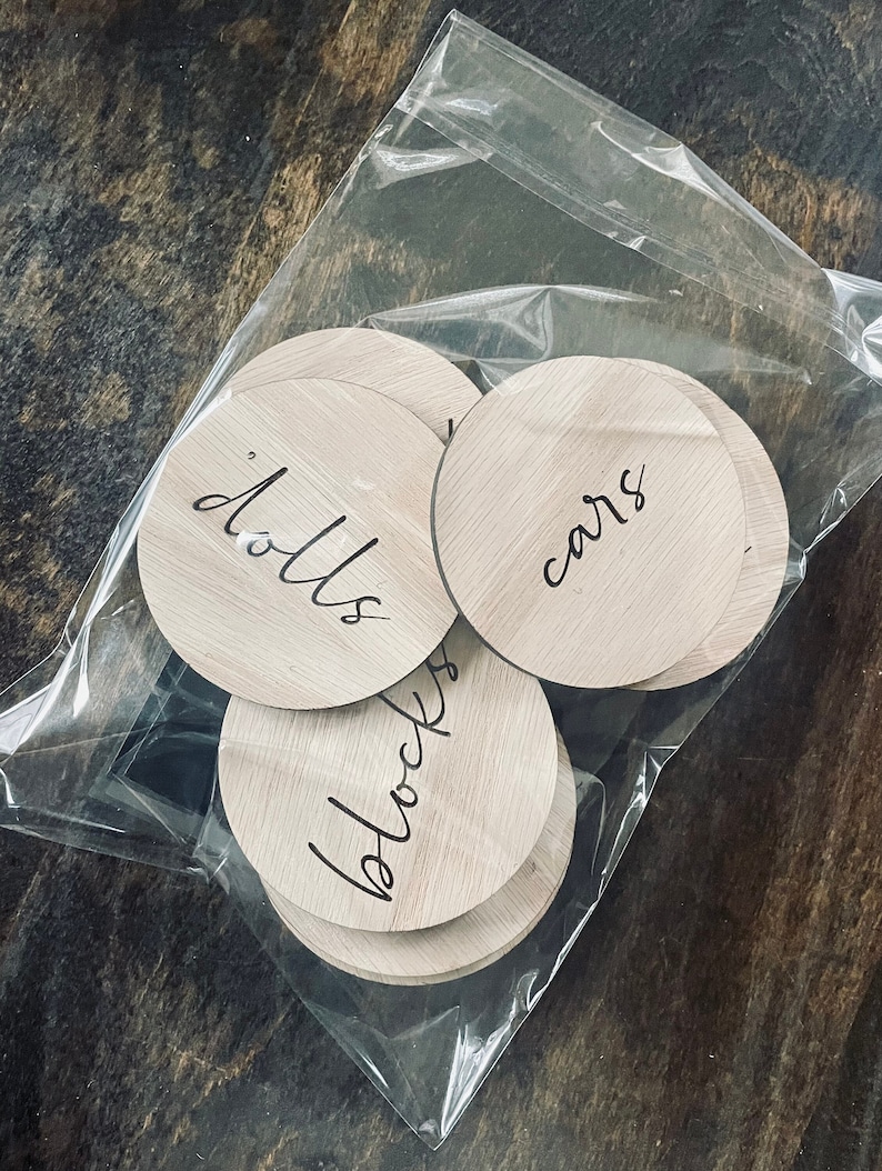 May include: A set of six wooden round labels with handwritten text. The labels are in a clear plastic bag. The labels say "dolls", "cars", "blocks", "books", "games", and "puzzles".