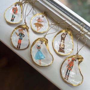 May include: Hand-painted oyster shell ornaments with gold trim and gold string hangers. The ornaments feature images of a nutcracker, a ballerina, and a grandfather clock. These unique ornaments are perfect for holiday decorating.