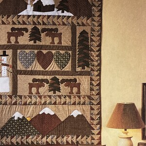 May include: A brown, beige, and green patchwork quilt with a winter theme. The quilt features a cabin, moose, trees, hearts, and mountains. The quilt is hanging on a wall.