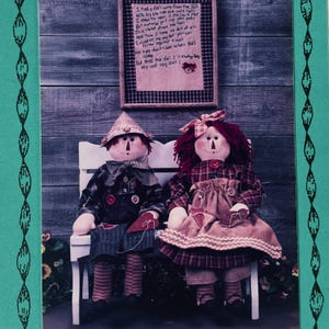 May include: Two fabric dolls sit on a white bench. The left doll wears a pointed hat and dark plaid clothing. The right doll has red yarn hair, a plaid dress, and a tan apron. Above them hangs a framed poem titled My Old Rag Doll.