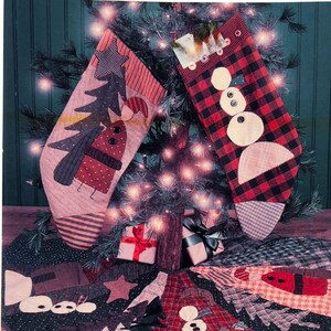 May include: Two fabric Christmas stockings and a tree skirt featuring patchwork designs. One stocking has a red and black buffalo check pattern with a snowman applique, while the other features a tree and bird motif. The tree skirt has matching patterns.