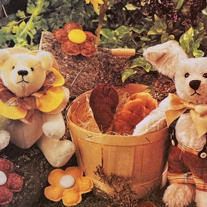May include: Two plush teddy bears, one with a yellow flower collar and the other with a brown bow tie, are sitting next to a basket filled with brown felt shapes. The bears are white with brown noses and eyes.