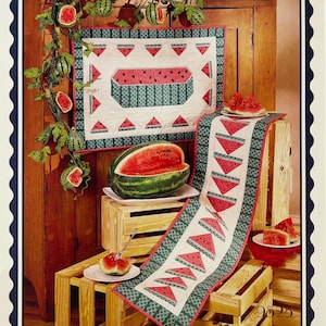 May include: A quilted wall hanging and a matching table runner featuring a watermelon motif with red fruit and green rind patterns on a cream background. A fresh watermelon half and slices are displayed on white plates near the textiles.