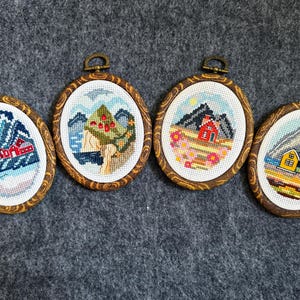 Può includere: Quattro opere d'arte ovali a punto croce raffiguranti scene di montagne e case. Ogni pezzo è incorniciato in un ovale di legno con un anello di metallo per appenderlo. Le palette di colori includono blu, rossi, gialli e verdi, che raffigurano paesaggi.