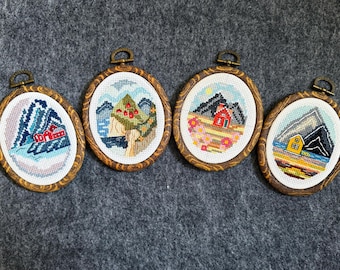Puede incluir: Cuatro obras de arte ovaladas de punto de cruz con escenas de montañas y casas. Cada pieza está enmarcada en un óvalo de madera con un lazo de metal para colgar. Las paletas de colores incluyen azules, rojos, amarillos y verdes, que representan paisajes.
