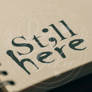 Könnte beinhalten: Nahaufnahme einer Seite mit den Worten "Still Here" in dunkelgrüner Schrift. Der Satz ist der Mittelpunkt, mit den Worten "Lof is Everywhere" in einem helleren Grün, die ein kreisförmiges Design um den Haupttext bilden.