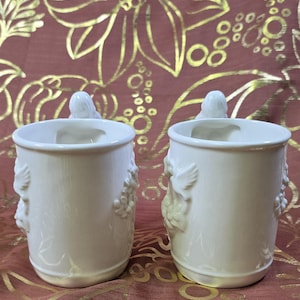 May include: Two off-white ceramic mugs with relief designs of birds and floral elements. Each mug has a bird-shaped handle and a decorative pattern of grapes and leaves. The mugs are set against a burgundy background with a gold floral pattern.