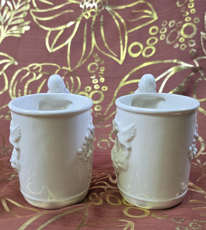May include: Two off-white ceramic mugs with relief designs of birds and floral elements. Each mug has a bird-shaped handle and a decorative pattern of grapes and leaves. The mugs are set against a burgundy background with a gold floral pattern.