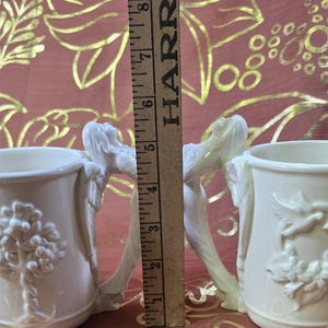 May include: Two off-white ceramic mugs with detailed relief designs. One mug features a tree, the other a floral wreath with a bird. The handles are sculpted figures. A wooden ruler shows the mugs are approximately 4 inches tall.