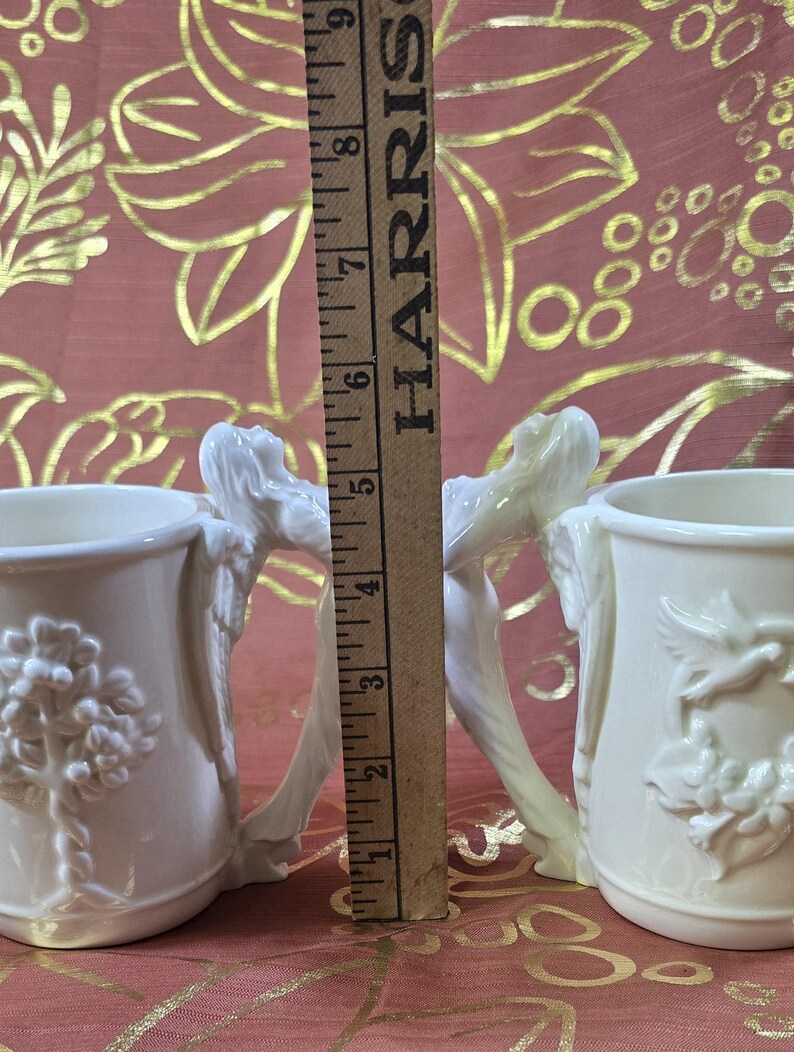 May include: Two off-white ceramic mugs with detailed relief designs. One mug features a tree, the other a floral wreath with a bird. The handles are sculpted figures. A wooden ruler shows the mugs are approximately 4 inches tall.