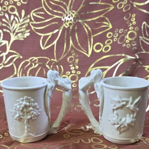 May include: Two off-white ceramic mugs. One mug features a tree design, the other a floral and bird design. Both mugs have handles shaped like angel figures. The mugs are set against a burgundy background with gold floral patterns.