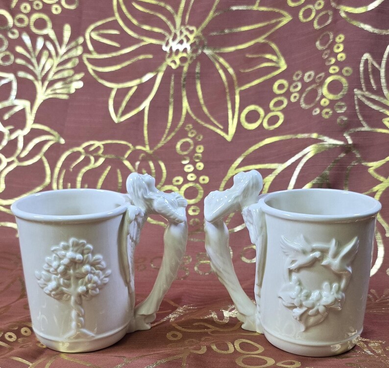 May include: Two off-white ceramic mugs. One mug features a tree design, the other a floral and bird design. Both mugs have handles shaped like angel figures. The mugs are set against a burgundy background with gold floral patterns.