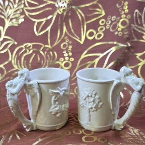 May include: Two off-white ceramic mugs with ornate handles and relief designs. One mug features a tree, the other a bird and floral motif. The handles are sculpted with a winged figure. The mugs are set against a burgundy background.