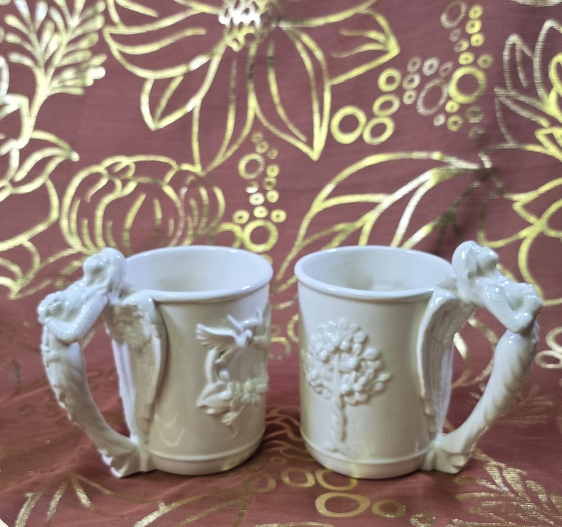 May include: Two off-white ceramic mugs with ornate handles and relief designs. One mug features a tree, the other a bird and floral motif. The handles are sculpted with a winged figure. The mugs are set against a burgundy background.
