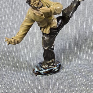 Puede incluir: Figura de cerámica de un hombre en pose de artes marciales. La figura viste una parte superior beige y pantalones negros, con una pierna levantada. La base es azul y marrón moteado. La figura mide unos 20 cm de altura.