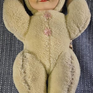 Può includere: Bambola di peluche vintage con viso dipinto e occhi chiusi. La bambola indossa un completo con cappuccio color crema con accenti floreali rosa. Le braccia e le gambe della bambola sono allungate, su un tessuto a righe blu e bianco.