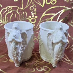 May include: Two white ceramic mugs with angel handles. The handles are sculpted in the form of an angel with outstretched wings and a rose. The mugs have a classic, elegant design, perfect for coffee or tea. The mugs are set against a patterned background.